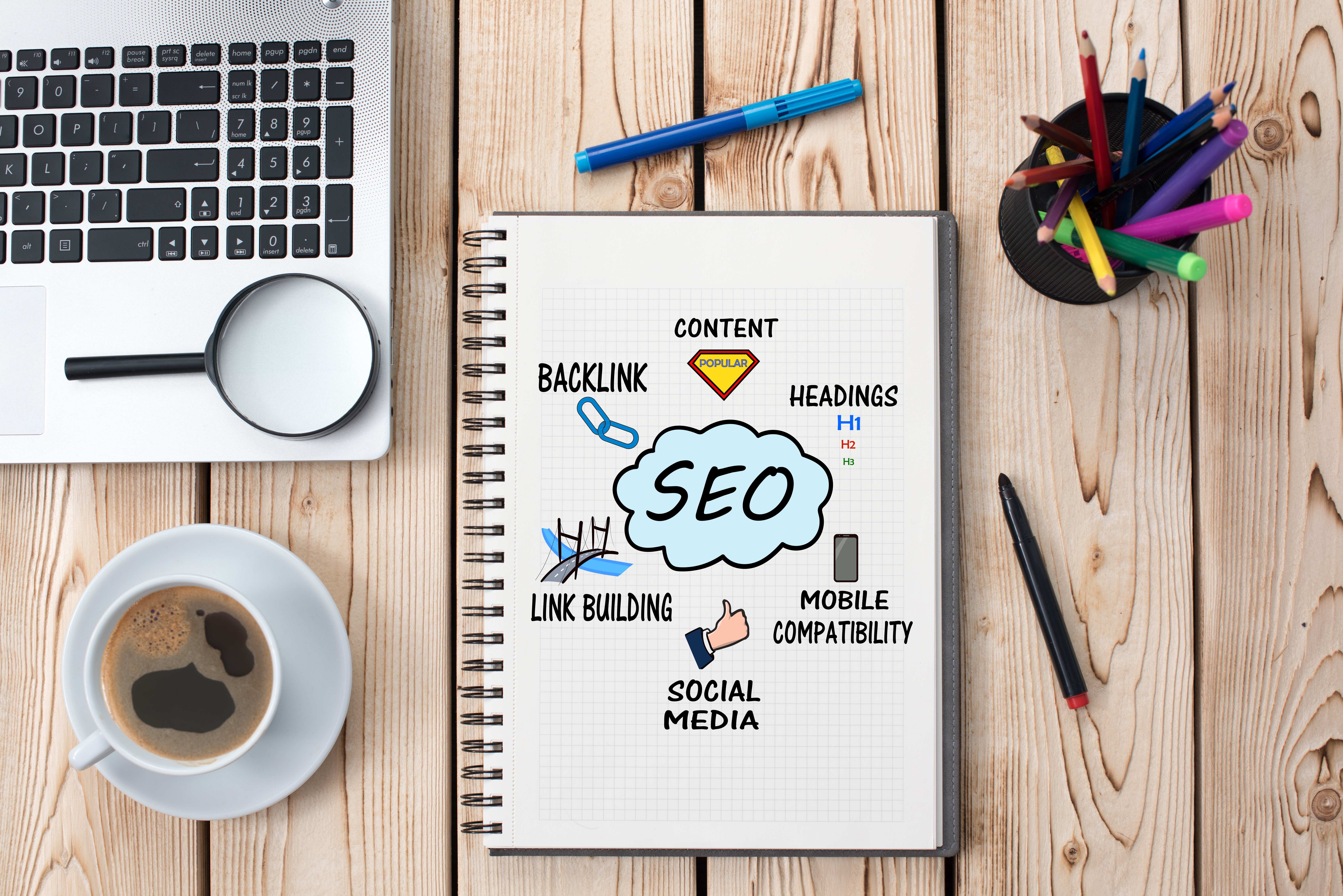 A notebook with SEO printed on its cover, a laptop, a cup of coffee, a magnifying glass, colorful pens and pencils in a black cup and a couple of pens all sitting on a table top or surface.